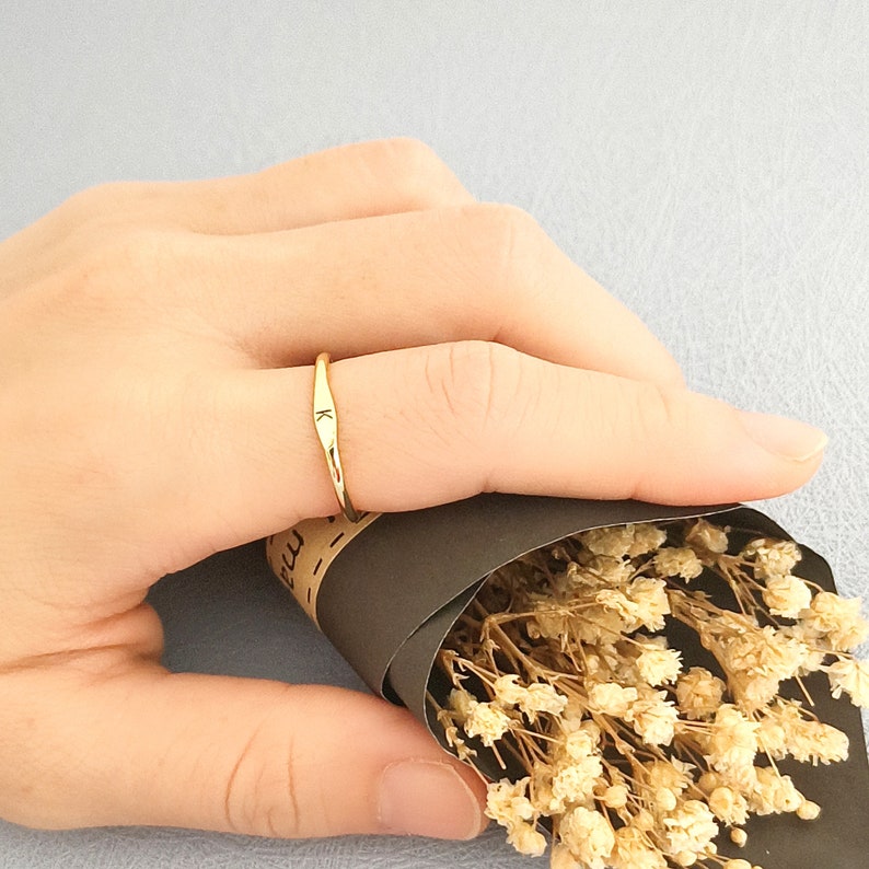 May include: A gold ring with a simple, hammered design and the letter "K" engraved on the band. The ring is worn on a person's finger.