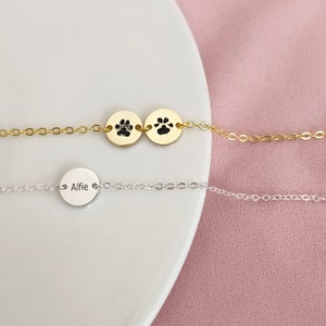 May include: Two gold necklaces with a paw print charm and a silver necklace with a round charm that says "Alfie" on a white surface.