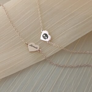 May include: Two gold heart necklaces, one with the word "Mummy" engraved on it and the other with two small hearts inside. The necklaces are on a beige fabric background.