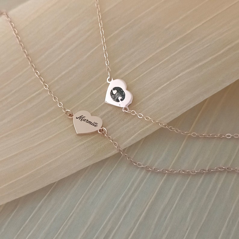 May include: Two gold heart necklaces, one with the word "Mummy" engraved on it and the other with two small hearts inside. The necklaces are on a beige fabric background.