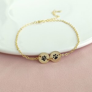 May include: A gold chain bracelet with two small round charms featuring paw prints. The charms are connected by a small gold chain.