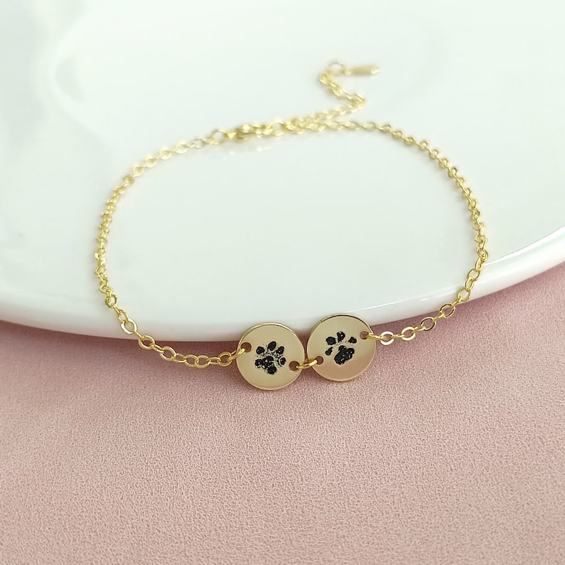 May include: A gold chain bracelet with two small round charms featuring paw prints. The charms are connected by a small gold chain.