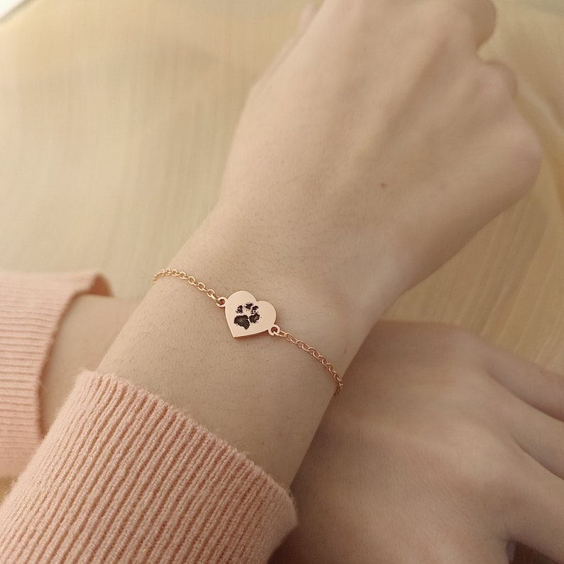 May include: A rose gold bracelet with a heart-shaped charm featuring a paw print. The bracelet is on a person's wrist.
