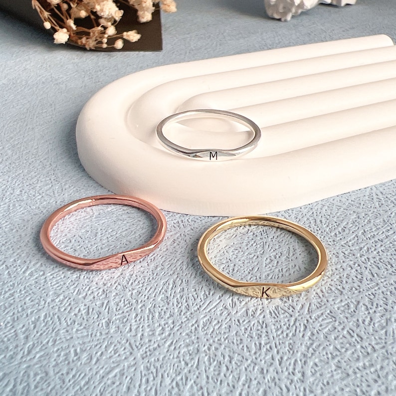 May include: Three simple, hammered metal rings with initial engravings. The rings are silver, rose gold, and gold. The initials are M, A, and K.