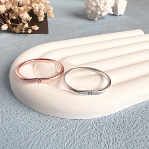 May include: Two simple, thin, personalized rings. One ring is rose gold with the letter "A" engraved on it. The other ring is silver with the letter "M" engraved on it.
