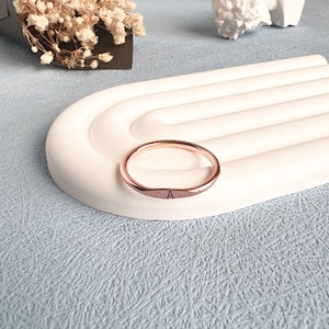 May include: A rose gold ring with the letter "A" engraved on the band. The ring is sitting on a white ceramic display stand.