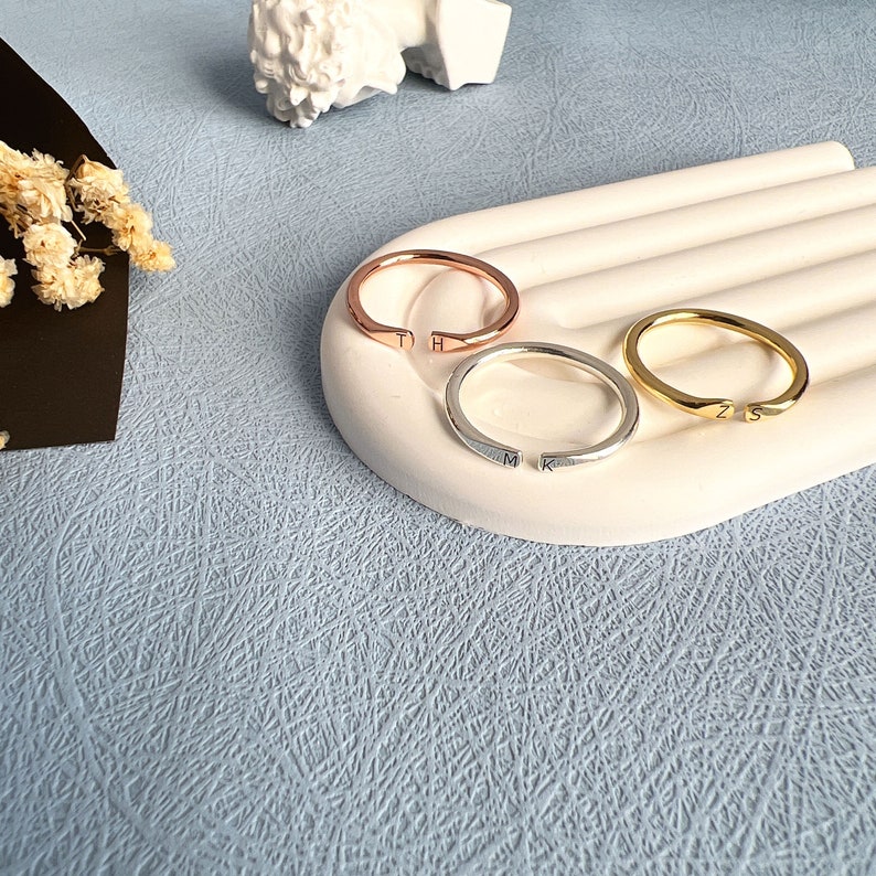 May include: Three simple gold rings with initials engraved on them. The rings are on a white ceramic surface. The rings are in rose gold, silver, and gold.