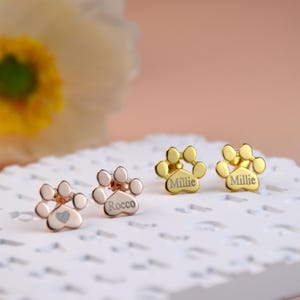 Op de afbeelding: Drie paar gouden poot afdruk oorbellen met gepersonaliseerde namen. De oorbellen liggen op een wit oppervlak. De namen op de oorbellen zijn Millie, Millie en Rocco.