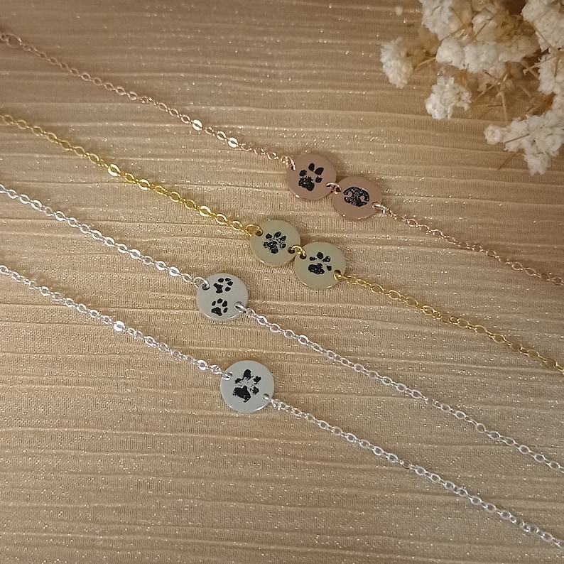 May include: Three silver chains with paw print charms. The chains are layered on a textured surface. The top chain is rose gold, the middle chain is gold, and the bottom chain is silver.