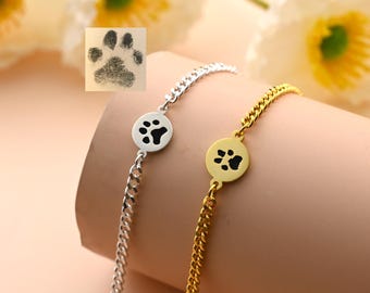 May include: Two bracelets, one silver and one gold, each featuring a circular charm with a black paw print design. The bracelets have chain links and are displayed on a neutral background with a floral accent.