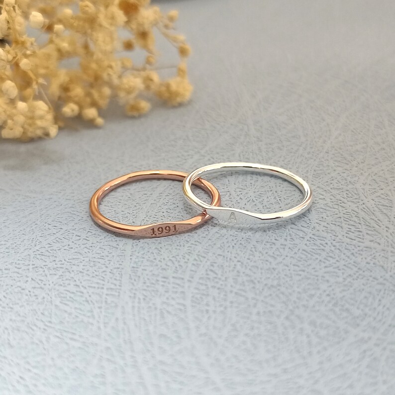 May include: Two thin, simple rings. One is silver and engraved with the letter "A". The other is rose gold and engraved with the year "1991".