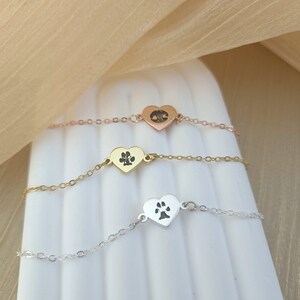 May include: Three delicate silver heart-shaped bracelets with a paw print design. The bracelets are layered on a white surface.