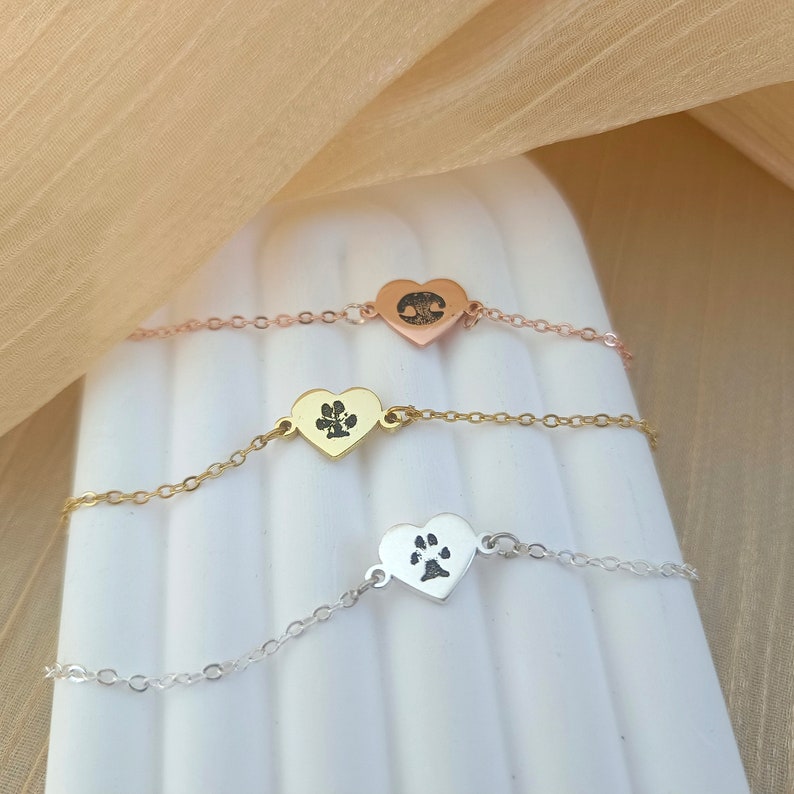May include: Three delicate silver heart-shaped bracelets with a paw print design. The bracelets are layered on a white surface.