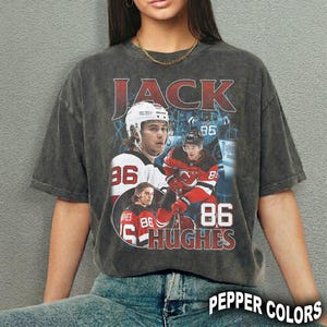 Puede incluir: Camiseta gris oscuro con un gráfico de hockey de estilo vintage. El diseño incluye el nombre "JACK", el número "86" y el nombre "HUGHES". El jugador de hockey está representado con uniforme.