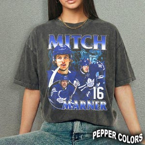 May include: Dark gray t-shirt featuring a graphic of hockey player Mitch Marner in a blue uniform. The design includes the name "Mitch Marner" and the number "16" in a stylized font. The shirt has a vintage, distressed look.