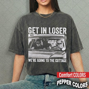 May include: A charcoal grey short-sleeve t-shirt with a vintage wash finish. The graphic features a car interior with two people wearing sunglasses, accompanied by the white text: GET IN LOSER WE'RE GOING TO THE COTTAGE printed in a bold font.