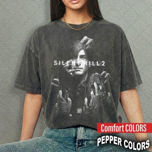 Puede incluir: Camiseta gris oscuro con un gráfico de Silent Hill 2. El diseño incluye un retrato con manos que se extienden hacia la cara y el texto "SILENT HILL 2". La camiseta es de la marca Comfort Colors.