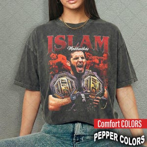 May include: Dark gray t-shirt featuring a graphic of Islam Makhachev holding two UFC championship belts. The word "ISLAM" is in red block letters above the image, with "Makhachev" in smaller white text. The shirt has "Comfort COLORS" and "PEPPER COLORS" printed on it.