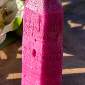 Puede incluir: Una punta de cristal rosa con una superficie rugosa y texturizada. El cristal es puntiagudo en la parte superior y tiene una base plana.