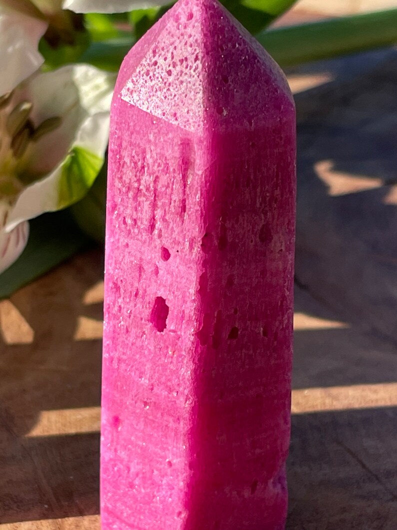 Puede incluir: Una punta de cristal rosa con una superficie rugosa y texturizada. El cristal es puntiagudo en la parte superior y tiene una base plana.