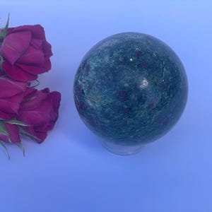 May include: A large, polished green sphere with red inclusions, resting on a clear stand. The sphere is next to a cluster of deep pink roses, creating a contrast of textures and colors. The overall theme is decorative.