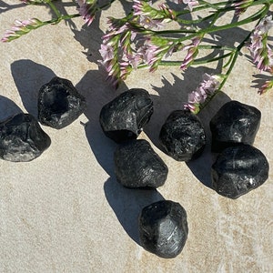 May include: A collection of eight black obsidian tumbled stones on a light beige surface. The stones are smooth and have a glossy finish.