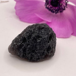 May include: A dark grey, rough, and textured rock with a small hole in the centre. The rock is sitting on a white surface.