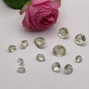 Puede incluir: Una colección de gemas verde claro de varias formas, incluyendo ovaladas, redondas y en forma de lágrima, se encuentran sobre una superficie blanca. Una rosa rosa con hojas verdes está en el fondo. Las gemas varían en tamaño.