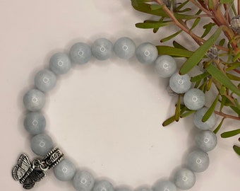 May include: A light blue beaded bracelet with a silver butterfly charm. The bracelet is made of round, smooth beads. The butterfly charm is detailed with etched wings. The bracelet is displayed on a white surface with green foliage.