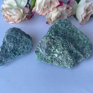 May include: Two rough, green mineral specimens with a sparkling texture are displayed. The stones are set against a light blue background, with several white and pink roses in the upper portion of the image.
