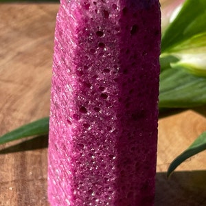 Puede incluir: Una punta de cristal de color morado oscuro con una superficie texturizada. El cristal tiene una punta puntiaguda y una base plana.