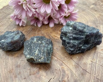 May include: Three rough, dark gray-blue crystals with textured surfaces are displayed on a wooden background. Soft pink flowers are arranged above the crystals. The crystals have a natural, organic appearance.