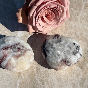 May include: Two polished, oval-shaped stones with white, gray, and burgundy patterns. A pink rose is positioned above them. The stones have a smooth, glossy texture, and the rose has multiple petals.