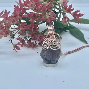 Puede incluir: Un colgante de piedra gris oscuro con un diseño de alambre de cobre, colgando de una cadena de plata.