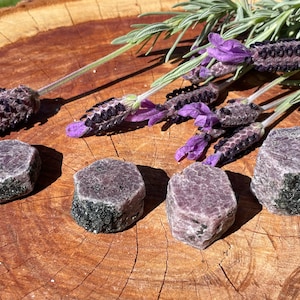 Puede incluir: Cinco cristales de rubí en bruto, de color púrpura oscuro y verde, sobre una superficie de madera. Los cristales son de forma hexagonal y tienen un aspecto natural y sin pulir.