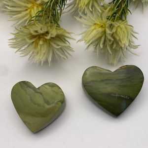 May include: Two heart-shaped stones with a green and brown marbled pattern. The stones are on a white surface, with a sprig of yellow flowers in the background. The stones are likely decorative or used in crafts.