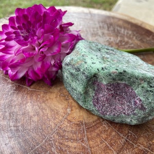 May include: A large, green and purple gemstone with a rough, natural texture. The stone is sitting on a wooden surface next to a pink and white flower.