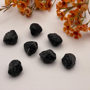 May include: Seven rough, black tourmaline stones are arranged on a white surface, with orange flowers in the background. The stones have a natural, unpolished appearance, with irregular shapes and textures. The arrangement suggests a focus on natural gemstones.