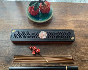 May include: A black and brown incense holder with a decorative floral design, a bundle of incense sticks, and a black incense stick holder. A small decorative floral branch and a small green dish with two red lychee fruits are also present.