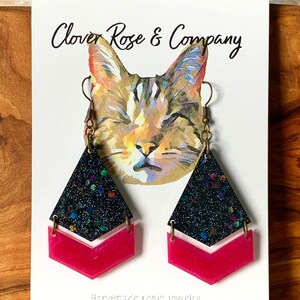K&ouml;nnte beinhalten: Ein Paar handgefertigte Ohrringe aus Harz mit einem schwarzen, schimmernden Glitzer-Oberteil und einem leuchtend pinkfarbenen Chevron-Unterteil. Die Ohrringe werden auf einer wei&szlig;en Karte mit dem Text "Clover Rose & Company" und "Handgefertigte Harzschmuck" pr&auml;sentiert.