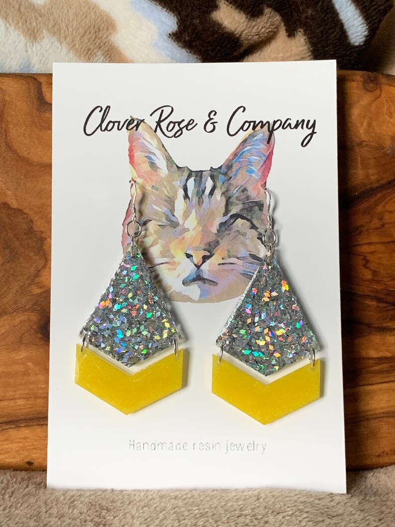 K&ouml;nnte beinhalten: Ein Paar handgefertigte Ohrringe aus Harz mit einem silbernen Glitzer-Chevron-Design und einem gelben Chevron-Akzent. Die Ohrringe werden auf einer wei&szlig;en Karte mit einer Katzenillustration und dem Text "Clover Rose & Company" und "Handgefertigte Harzohrringe" gezeigt.