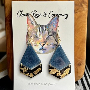 K&ouml;nnte beinhalten: Ein Paar handgefertigte Resin-Ohrringe mit einem geometrischen Design in Blau und Gold. Die Ohrringe sind tropfenf&ouml;rmig und haben einen goldenen Akzent. Die Ohrringe werden auf einer Karte mit einer Katzenillustration und dem Text "Clover Rose & Company" und "Handgefertigte Resin-Schmuckst&uuml;cke" pr&auml;sentiert.