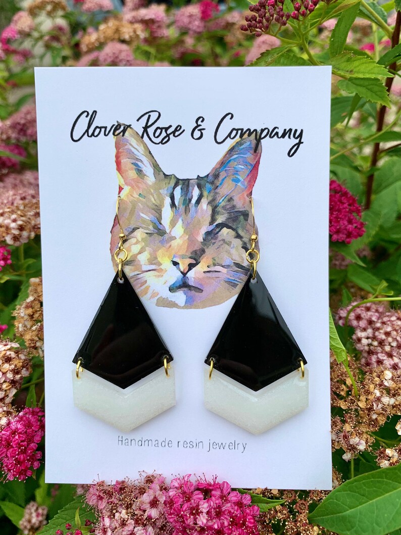 K&ouml;nnte beinhalten: Schwarze und wei&szlig;e geometrische Harzohrringe mit goldenen Haken. Die Ohrringe werden auf einer wei&szlig;en Karte mit dem Text "Clover Rose & Company" und "Handgefertigte Harzschmuck" pr&auml;sentiert.