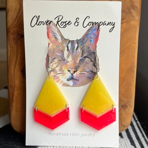 K&ouml;nnte beinhalten: Ein Paar handgefertigte Resin-Ohrringe mit einem gelben und pinkfarbenen Chevron-Muster. Die Ohrringe werden auf einer Karte mit einer Katzenillustration und dem Text "Clover Rose & Company" und "Handgefertigte Resin-Schmuckst&uuml;cke" pr&auml;sentiert.