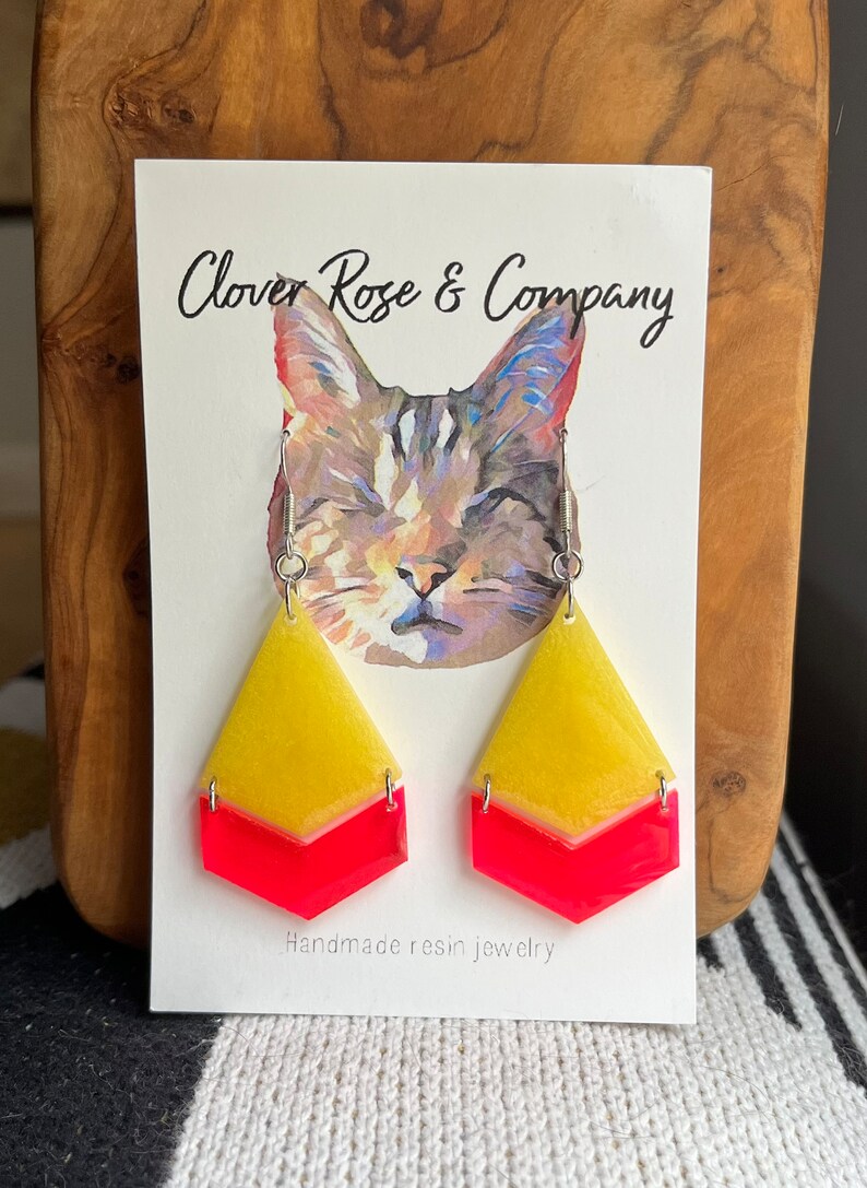 K&ouml;nnte beinhalten: Ein Paar handgefertigte Resin-Ohrringe mit einem gelben und pinkfarbenen Chevron-Muster. Die Ohrringe werden auf einer Karte mit einer Katzenillustration und dem Text "Clover Rose & Company" und "Handgefertigte Resin-Schmuckst&uuml;cke" pr&auml;sentiert.