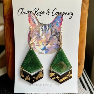 K&ouml;nnte beinhalten: Gr&uuml;ne und goldene Harzohrringe in geometrischer Form. Die Ohrringe werden auf einer Karte mit einer Aquarell-Illustration einer Katze und dem Text "Clover Rose & Company" und "Handgefertigte Harzschmuck" pr&auml;sentiert.