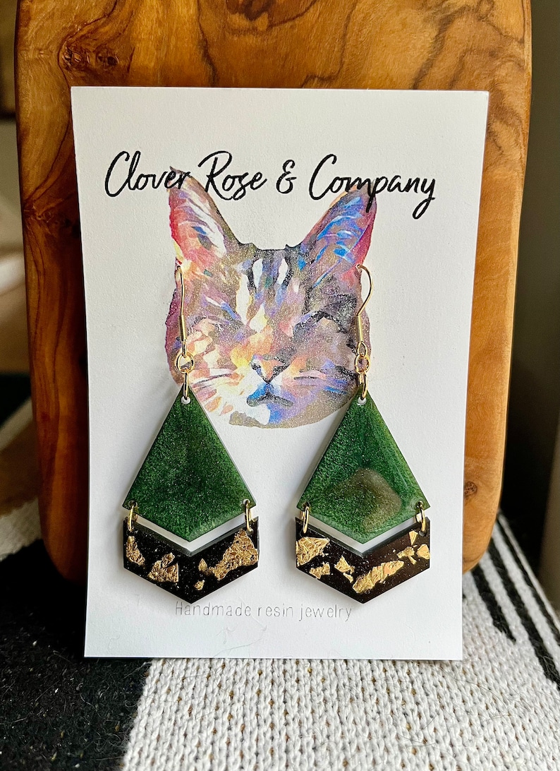 K&ouml;nnte beinhalten: Gr&uuml;ne und goldene Harzohrringe in geometrischer Form. Die Ohrringe werden auf einer Karte mit einer Aquarell-Illustration einer Katze und dem Text "Clover Rose & Company" und "Handgefertigte Harzschmuck" pr&auml;sentiert.