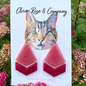 K&ouml;nnte beinhalten: Ein Paar rosafarbene und wei&szlig;e Ohrringe aus Harz mit geometrischem Design. Die Ohrringe befinden sich auf einer wei&szlig;en Karte mit dem Text "Clover Rose & Company" und "Handgefertigte Harzschmuck".