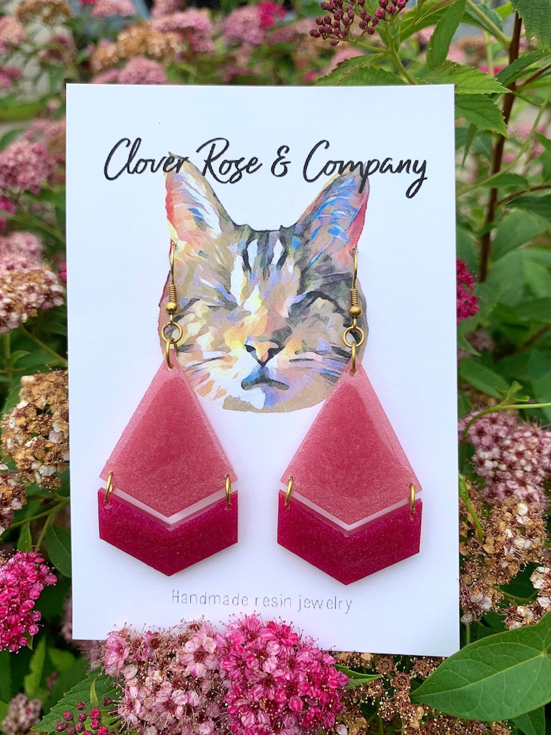 K&ouml;nnte beinhalten: Ein Paar rosafarbene und wei&szlig;e Ohrringe aus Harz mit geometrischem Design. Die Ohrringe befinden sich auf einer wei&szlig;en Karte mit dem Text "Clover Rose & Company" und "Handgefertigte Harzschmuck".