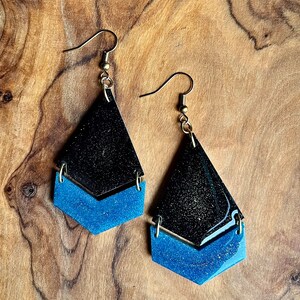 K&ouml;nnte beinhalten: Ein Paar Ohrh&auml;nger mit schwarzem und blauem Glitzer-Finish. Die Ohrringe sind tropfenf&ouml;rmig mit einer kleineren Tropfenform darunter.